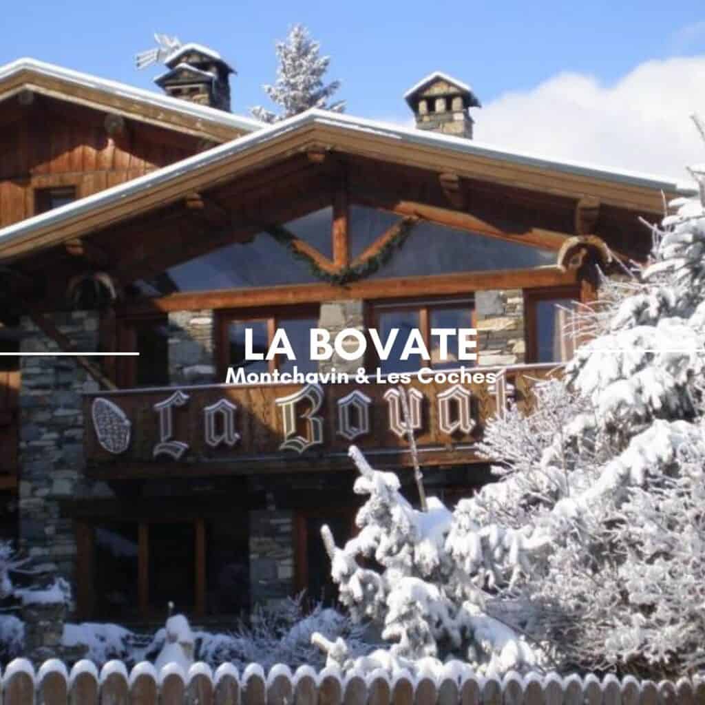 A traditional alpine chalet with a snow-covered roof and a wooden sign reading "La Bovate." The stone and wood exterior, along with the surrounding snow-laden trees, create a picturesque winter scene. The text overlay reads "La Bovate, Montchavin & Les Coches."