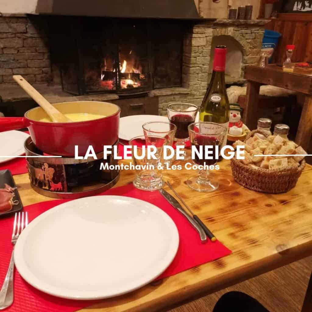 A rustic wooden dining table set for a cozy fondue meal, featuring a red pot of melted cheese, plates, glasses of red wine, and a basket of bread cubes. A crackling stone fireplace in the background adds to the warm alpine atmosphere. The text overlay reads "La Fleur de Neige, Montchavin & Les Coches.