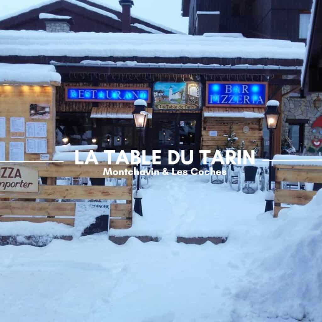 A charming snow-covered restaurant entrance with glowing blue signs for the bar, pizzeria, and dining area. The outdoor seating is partially buried in fresh snow, enhancing the cozy winter atmosphere. The text overlay reads "La Table du Tarin, Montchavin & Les Coches."