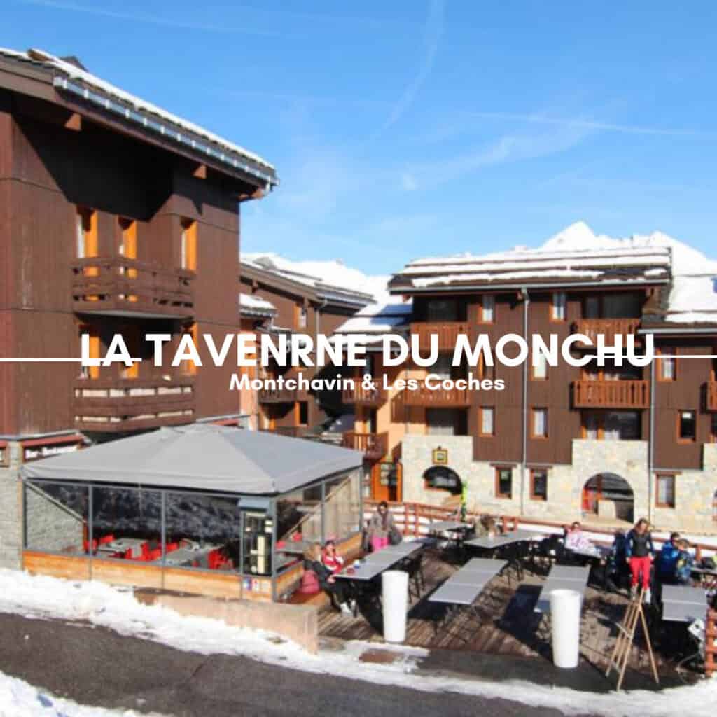 A lively outdoor dining area at La Taverne du Monchu, a chalet-style restaurant in Montchavin & Les Coches. Wooden buildings with snow-covered roofs surround the terrace, where guests enjoy meals and drinks under a clear blue sky. A partially enclosed seating area with red chairs provides a cozy spot for winter dining.