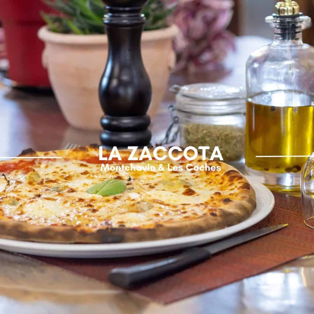 A wood-fired pizza with a golden crust and melted cheese, garnished with a fresh basil leaf, sits on a wooden table. Olive oil, spices, and a pepper grinder complete the rustic dining setup. The text overlay reads "La Zaccota, Montchavin & Les Coches.