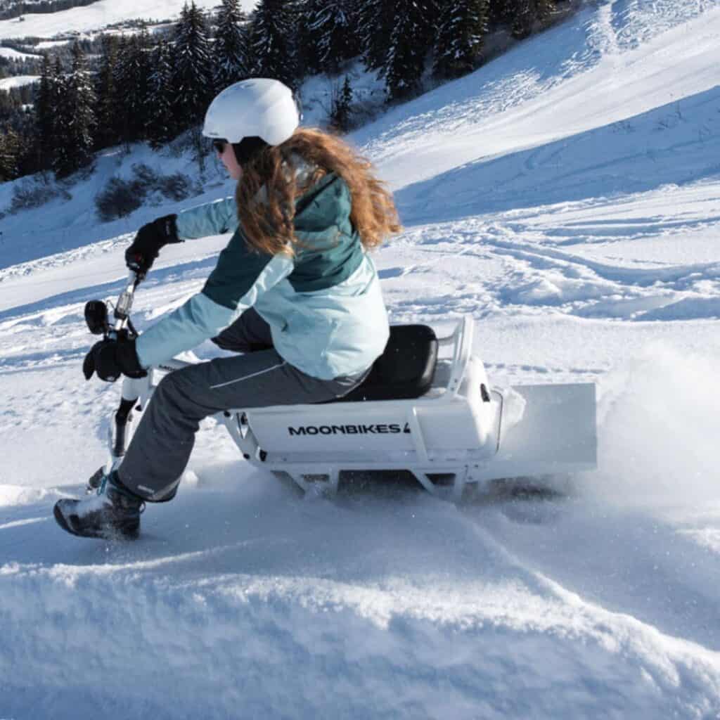 A person in winter gear and a helmet rides a MoonBike across a snowy slope in La Plagne, kicking up powder as they navigate the terrain. The electric snow bike, labeled "MoonBikes," glides effortlessly over the fresh snow with a scenic mountain backdrop.
