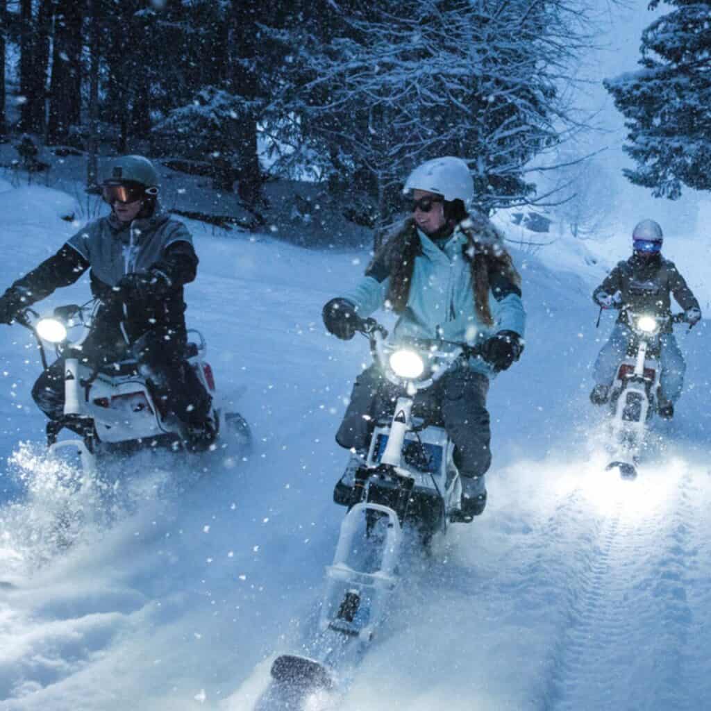 A group of moonbikers rides through a snowy forest in La Plagne at dusk, their headlights illuminating the falling snow. The riders, dressed in winter gear and helmets, carve a path through the deep powder on their MoonBikes, enjoying an exhilarating nighttime adventure.