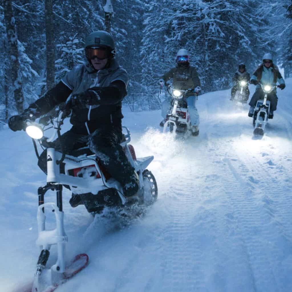A leading rider speeds down a snow-covered trail in La Plagne on a MoonBike, followed closely by a group of fellow moonbikers. Their headlights cut through the snowy evening, lighting up the path as they explore the winter landscape.