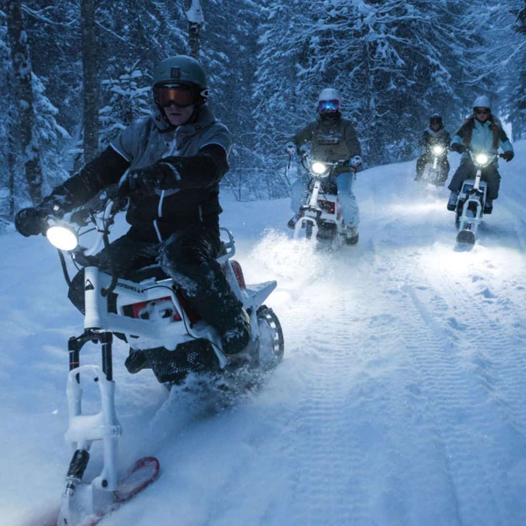 A leading rider speeds down a snow-covered trail in La Plagne on a MoonBike, followed closely by a group of fellow moonbikers. Their headlights cut through the snowy evening, lighting up the path as they explore the winter landscape.