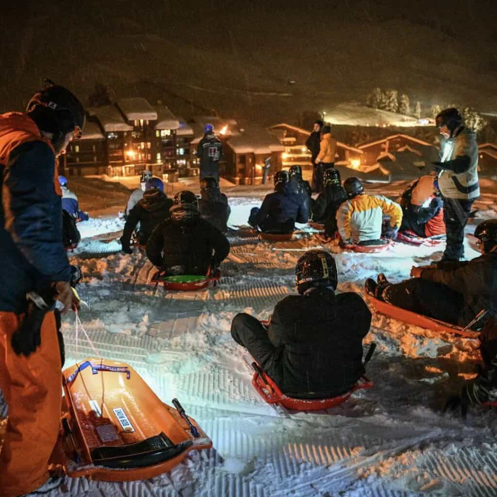 A group of people in helmets and winter gear sit on sleds at the top of a snowy hill, preparing for a night-time sledding adventure in La Plagne. The scene is illuminated by headlamps and nearby chalet lights, creating a cozy contrast against the dark, snowy mountain landscape.