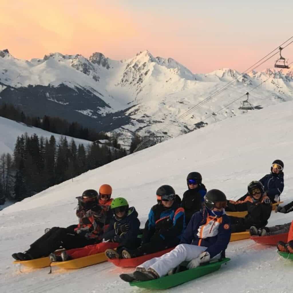 A group of sledders, dressed in colorful winter gear and helmets, sit on sleds atop a snow-covered slope in La Plagne, with the stunning backdrop of the French Alps at sunset. The ski lifts and distant peaks add to the picturesque winter adventure setting.