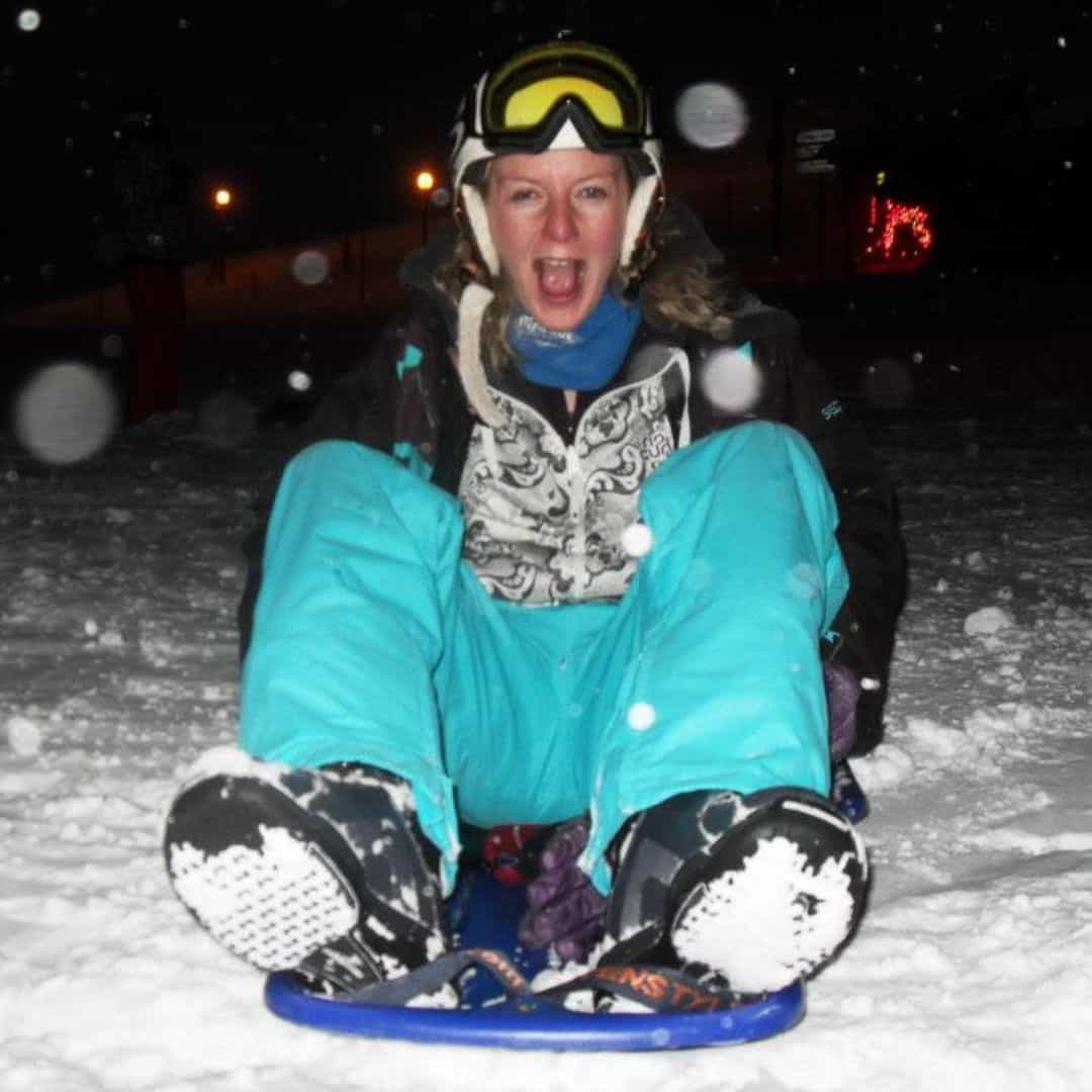 A woman in bright blue snow pants and a patterned jacket excitedly rides a sled down a snowy hill during a night-time sledding experience in La Plagne. Snowflakes fill the air as she grins with excitement, wearing ski goggles pushed up onto her helmet.