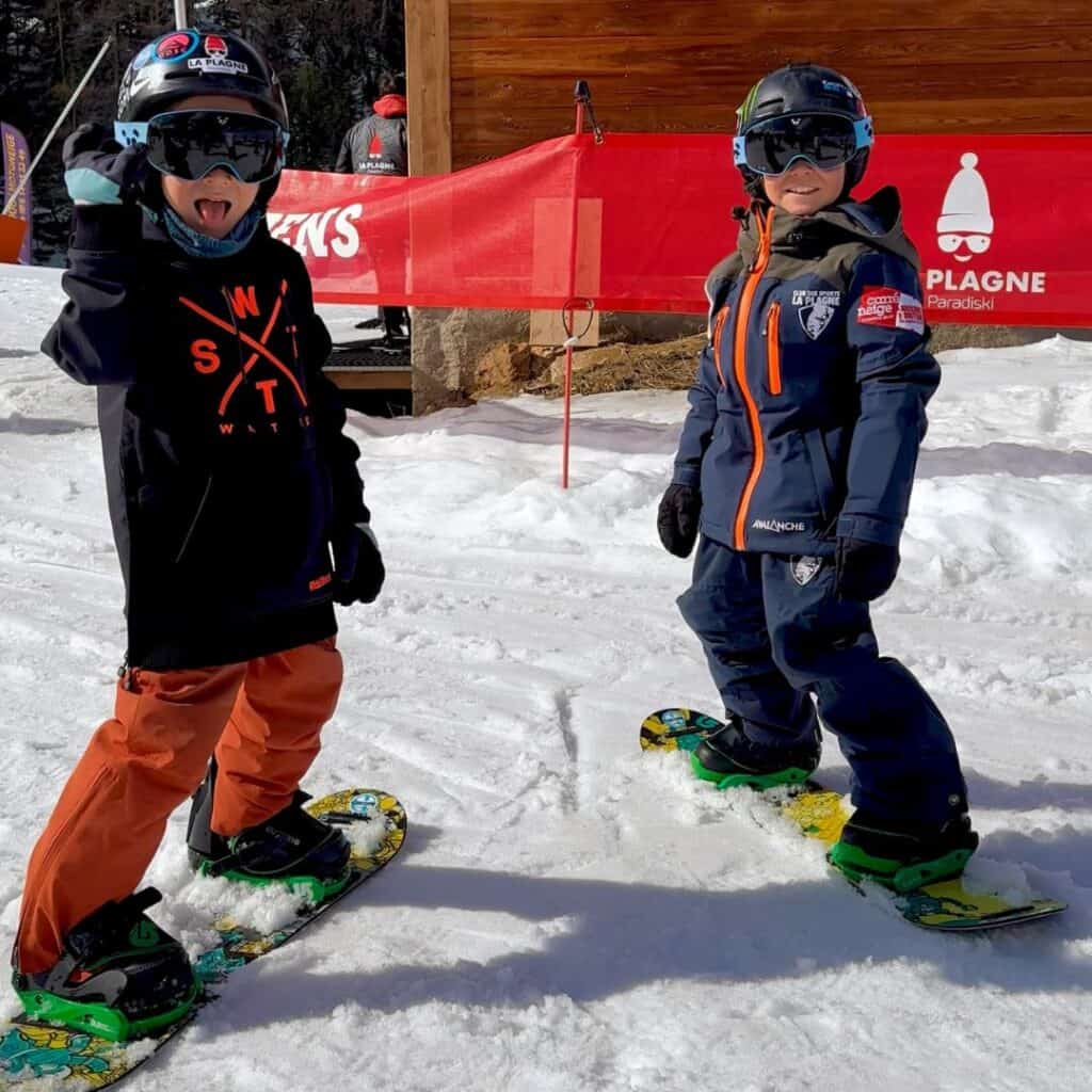 Two kids in full snowboarding gear smile and pose on the slopes at La Plagne, one making a playful face. A wooden lodge and red “La Plagne Paradiski” signage sit behind them.