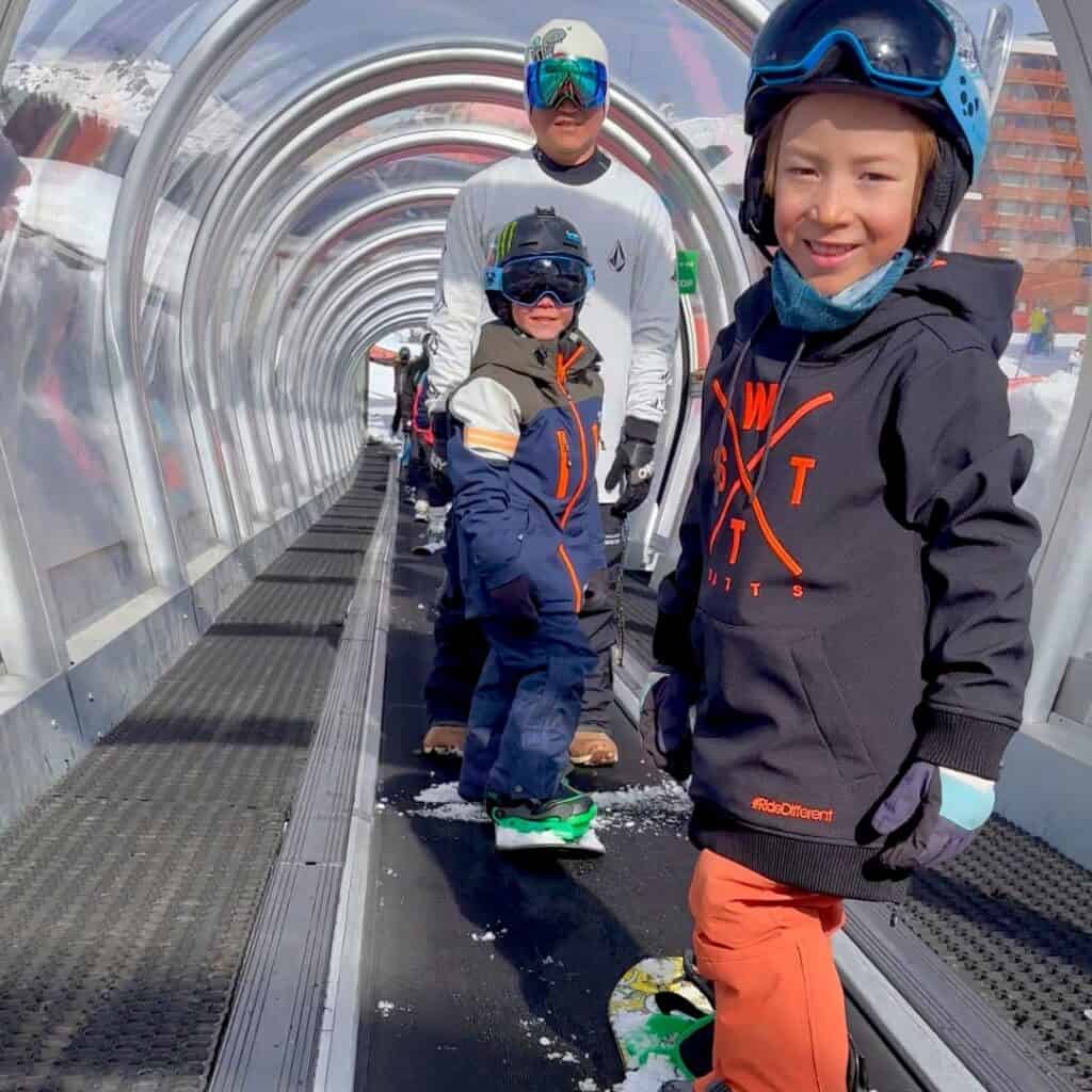 Two young snowboarders, supervised by an adult, ride a magic carpet lift inside a transparent tunnel in La Plagne, France. All wearing helmets and snow gear, the kids demonstrate key elements of snowboarding safety for kids in La Plagne, including proper supervision and protective equipment.