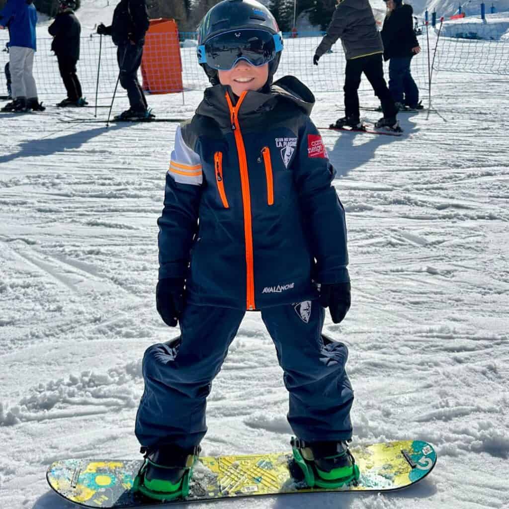A Smiling young child in full snowboarding gear stands confidently on a snowboard in the snow, wearing a helmet, goggles, and a navy snowsuit marked “Club de Sports La Plagne.” This cheerful image supports the topic of what age to start snowboarding by showcasing a young beginner on the slopes.