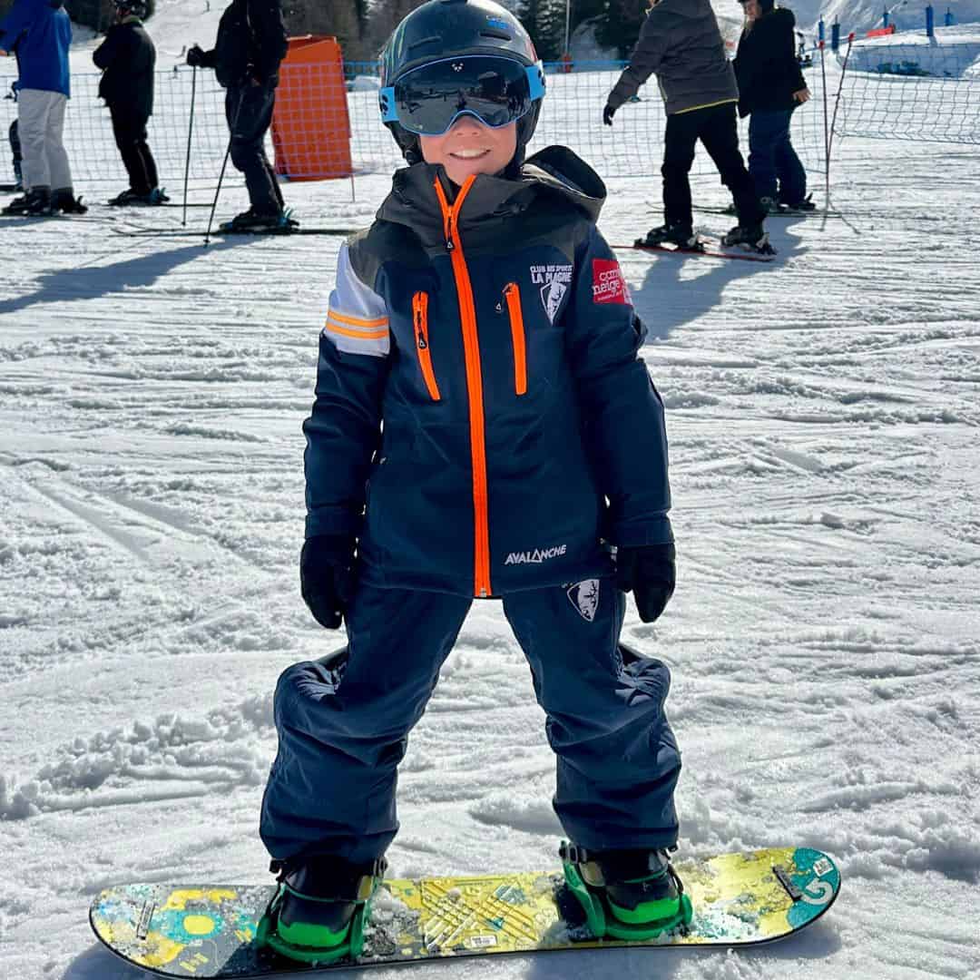 A Smiling young child in full snowboarding gear stands confidently on a snowboard in the snow, wearing a helmet, goggles, and a navy snowsuit marked “Club de Sports La Plagne.” This cheerful image supports the topic of what age to start snowboarding by showcasing a young beginner on the slopes.