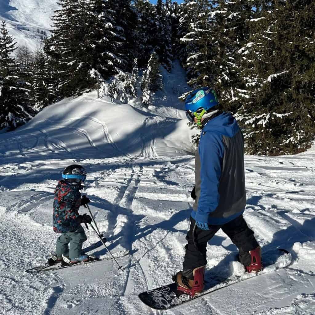 An adult snowboarder and a young skier head down a snowy forest trail in La Plagne, capturing the versatility of the resort for anyone deciding between skiing or snowboarding in La Plagne.