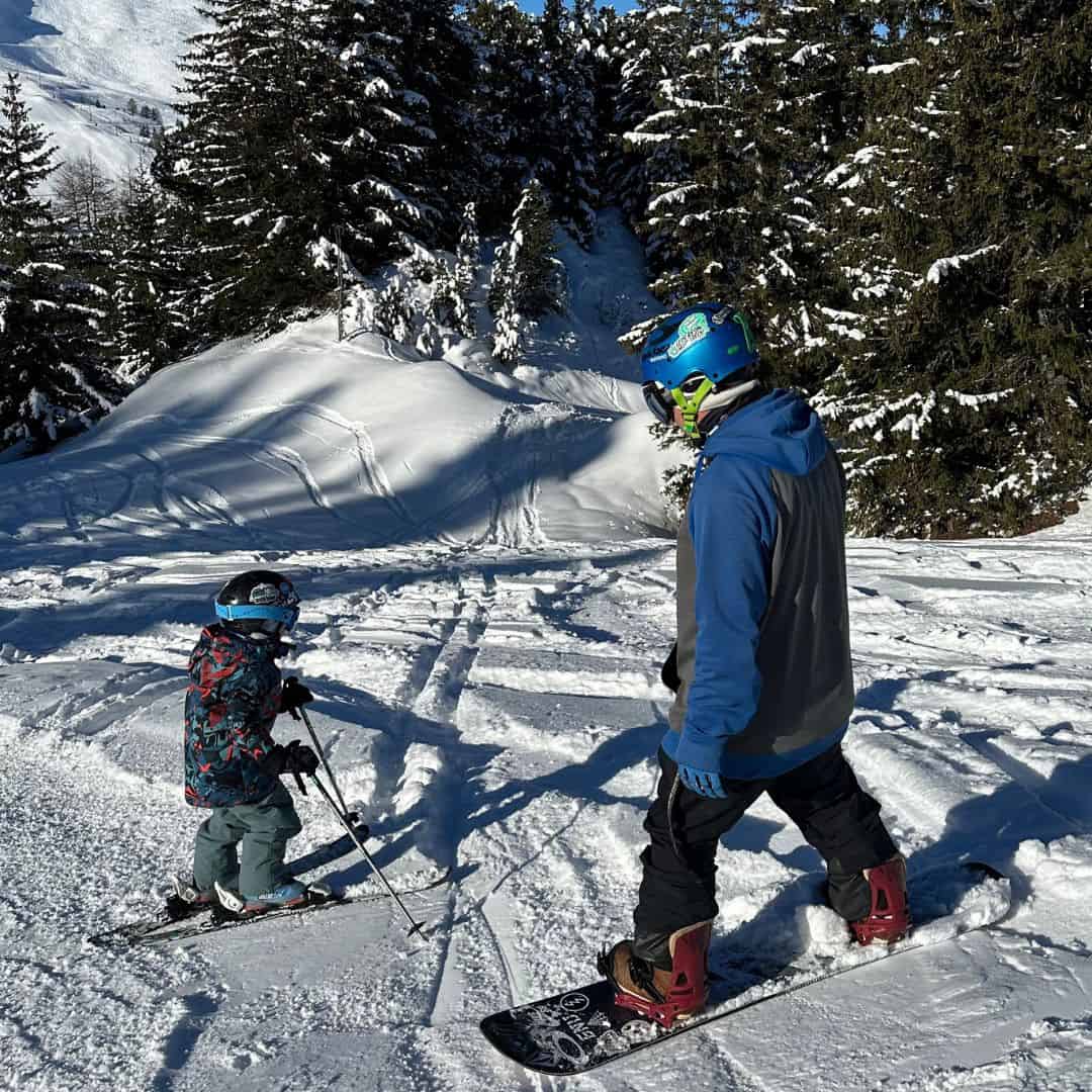 An adult snowboarder and a young skier head down a snowy forest trail in La Plagne, capturing the versatility of the resort for anyone deciding between skiing or snowboarding in La Plagne.