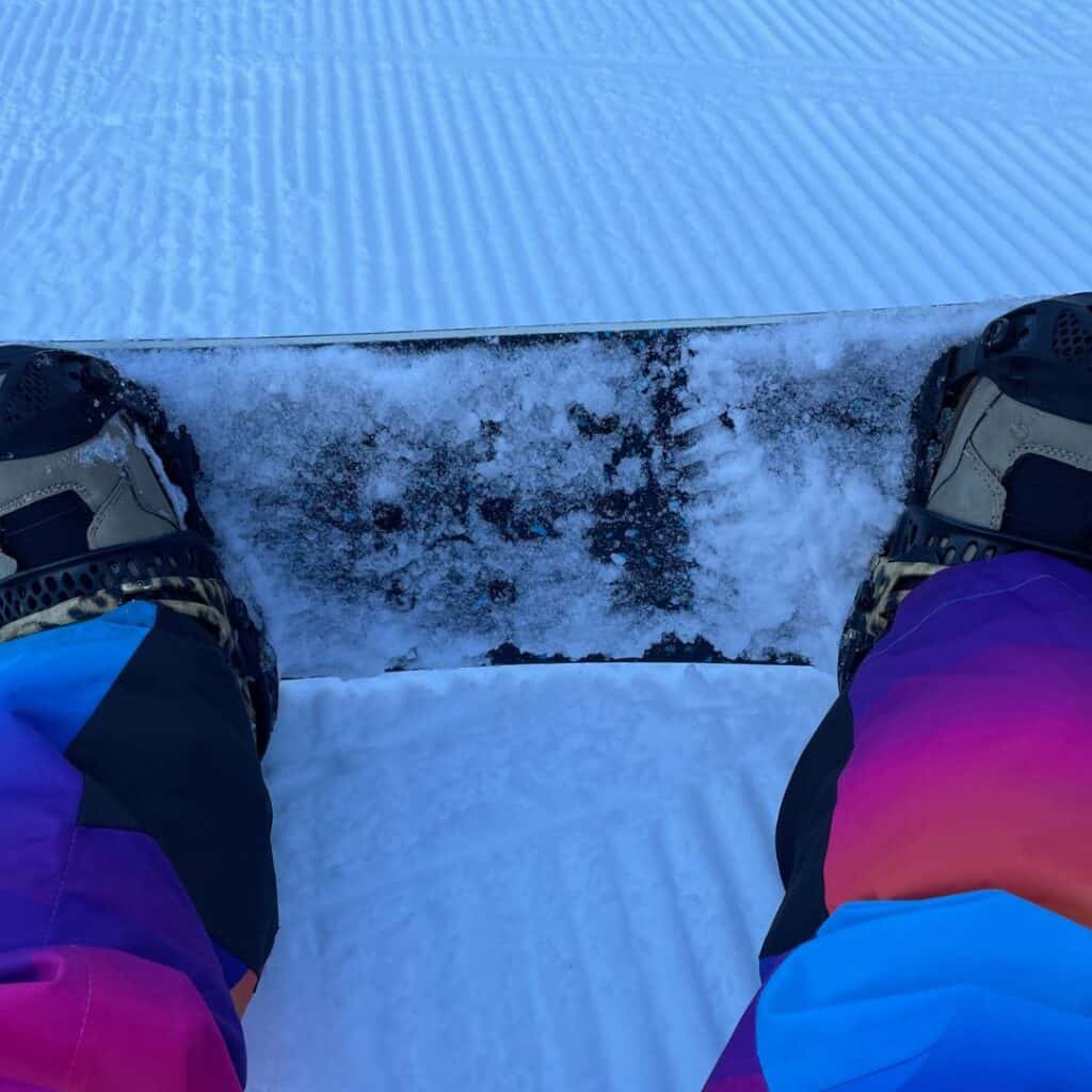 Looking down at a snowboard strapped in on a freshly groomed slope, with bold-colored pants in view—this shot captures the thrill of choosing skiing or snowboarding in La Plagne first thing in the morning.