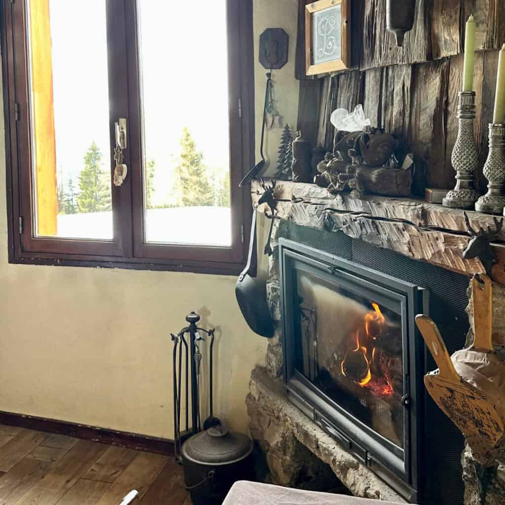 A cozy fireplace corner at Chalet du Plan Bois, featuring a crackling fire, rustic decor, and a window view of snowy alpine trees outside.