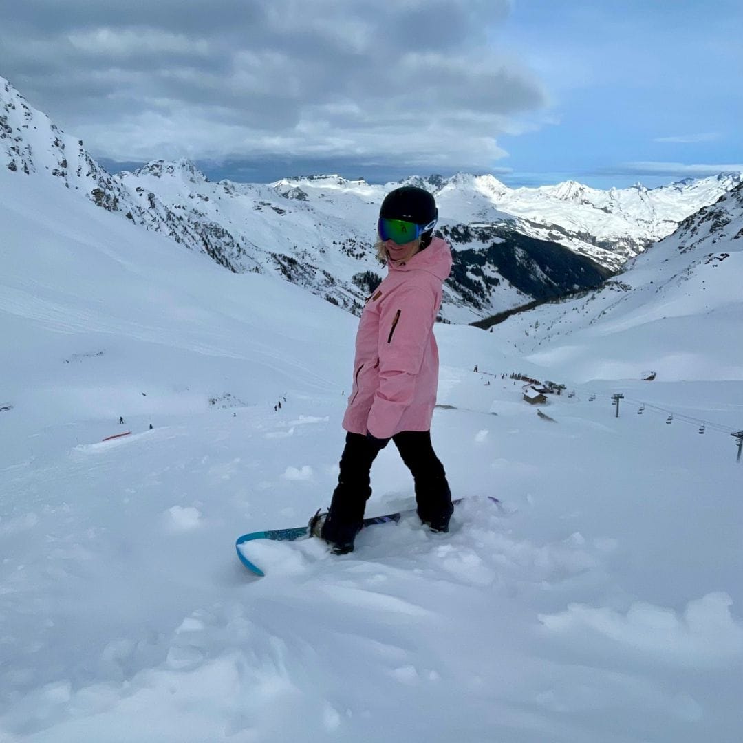 A snowboarder in a pink jacket smiles while standing on a snowy ridge, surrounded by stunning alpine peaks at La Plagne—ideal scenery for skiing or snowboarding adventures.