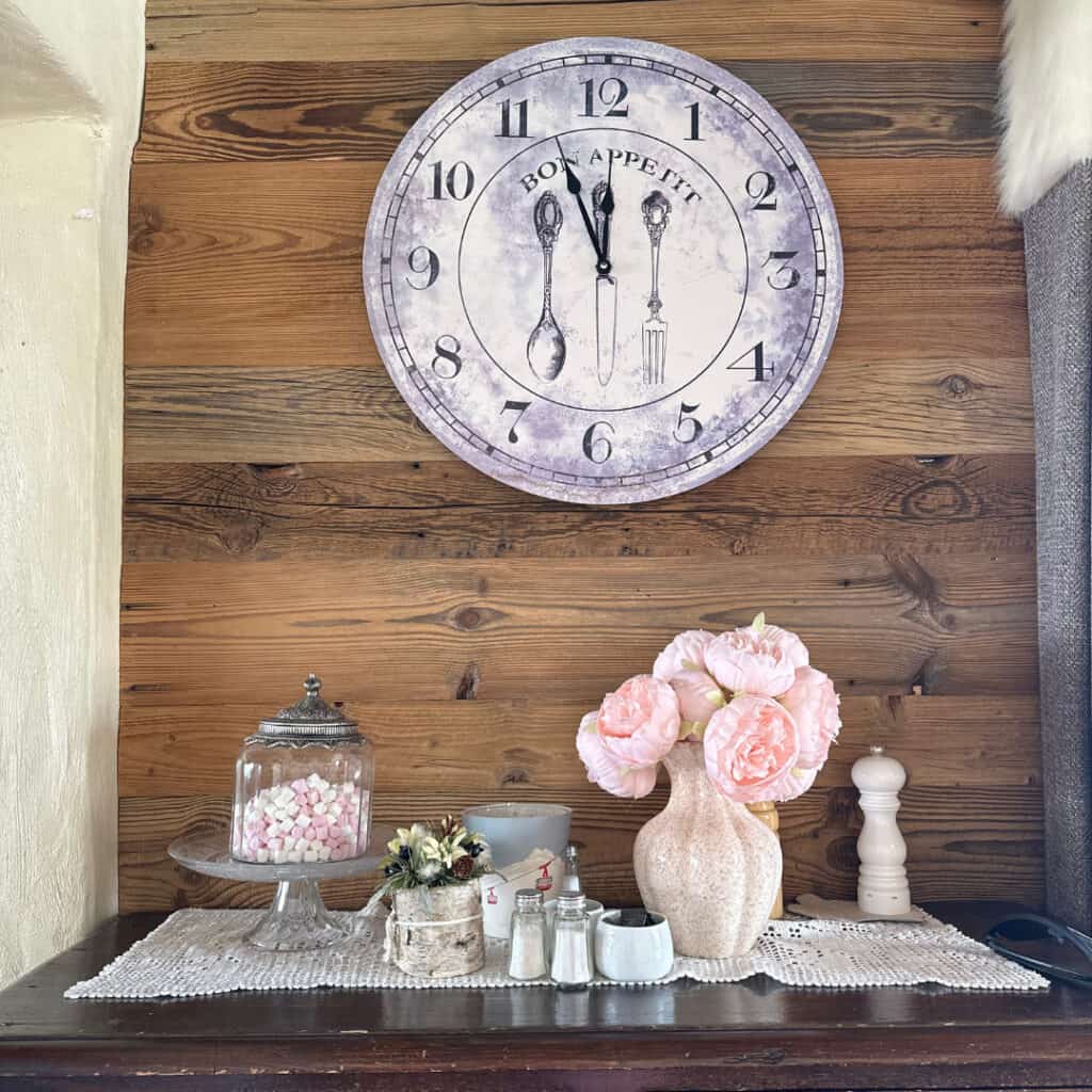 A quaint setup at Chalet du Plan Bois with a “Bon Appétit” wall clock, a soft pink flower arrangement, jars of sugar and spices, and a glass jar of mini marshmallows on a lace runner.