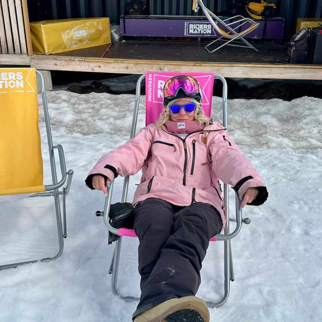 A woman in a pink snow jacket relaxes in a lounge chair labeled “Riders Nation” on a snowy patio, showing off the chill side of skiing or snowboarding in La Plagne.