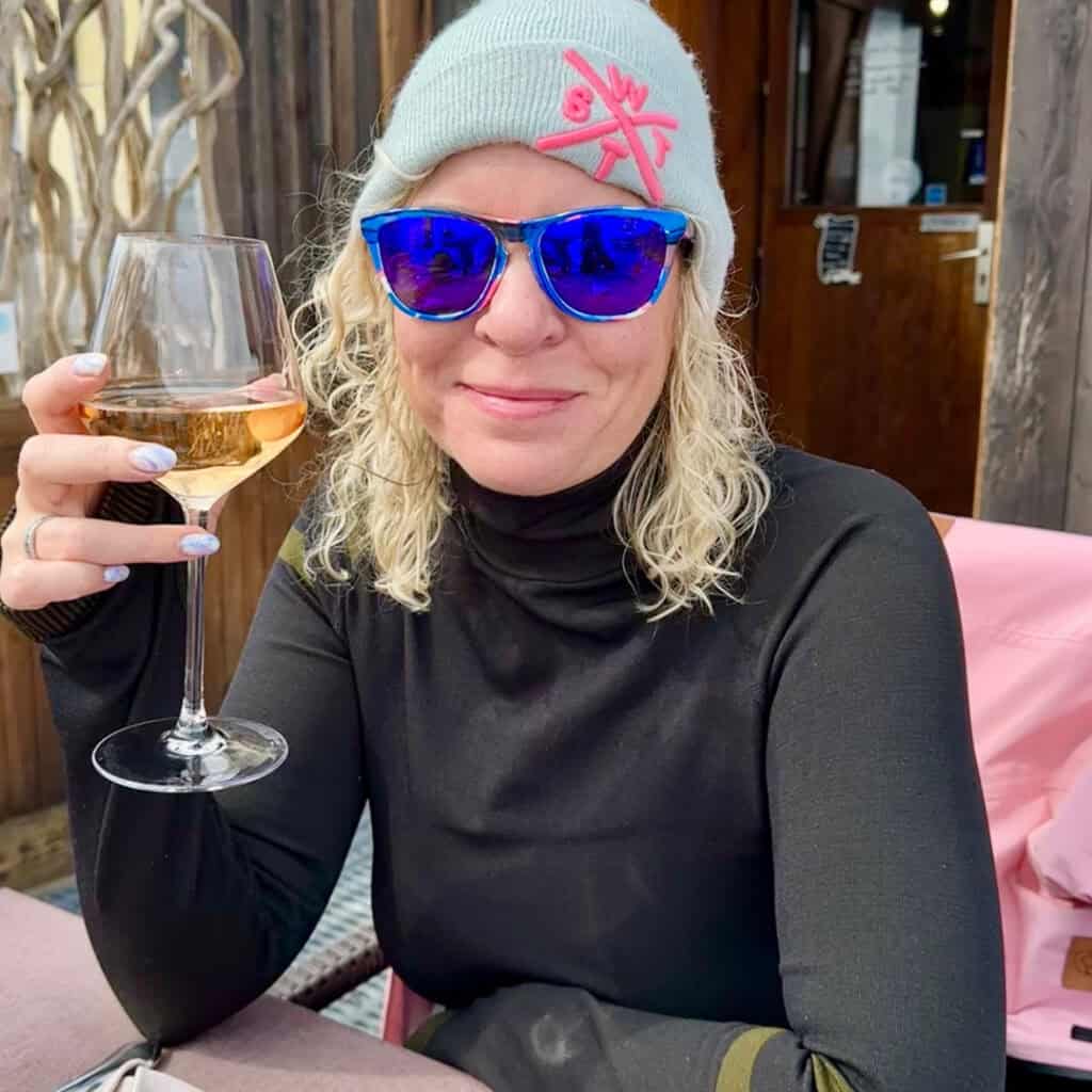 A woman in a light blue beanie and mirrored sunglasses smiles while enjoying a glass of rosé wine on the terrace of Chalet du Plan Bois, dressed warmly for the alpine chill.