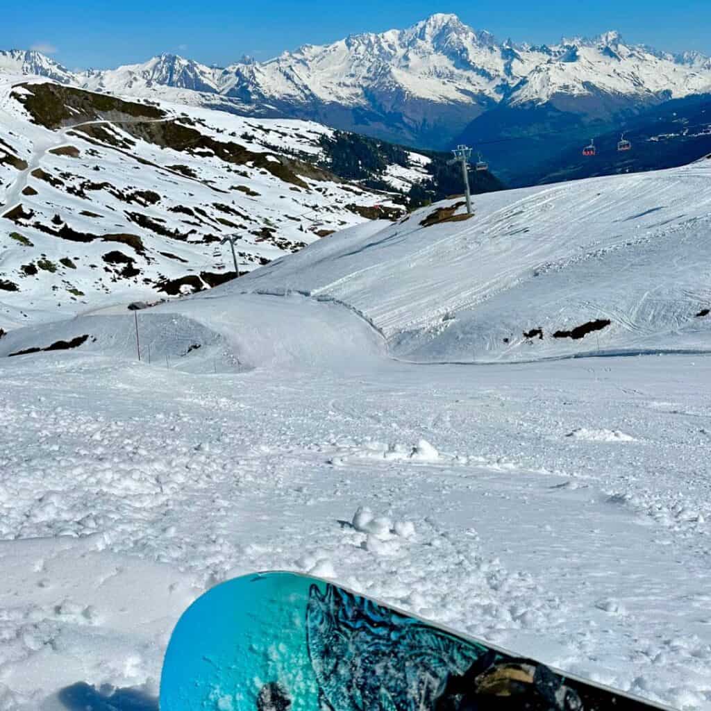 A snowboarder sits at the top of a broad slope in La Plagne with a clear view of the surrounding mountains, highlighting the epic terrain available for both skiing or snowboarding in La Plagne.