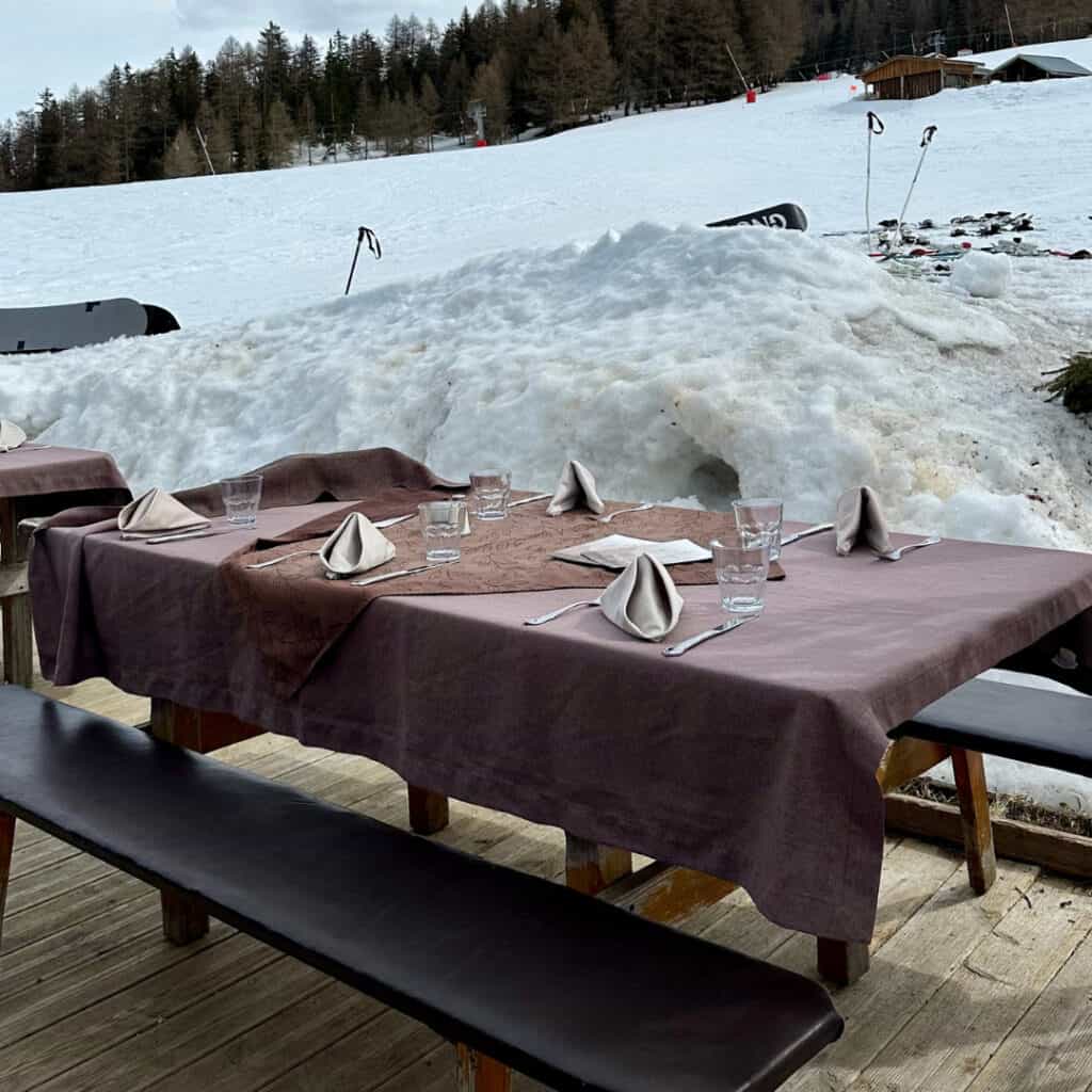 An outdoor dining setup at Chalet du Plan Bois with rustic wooden tables draped in brown tablecloths, set for service against a snowy mountain backdrop.
