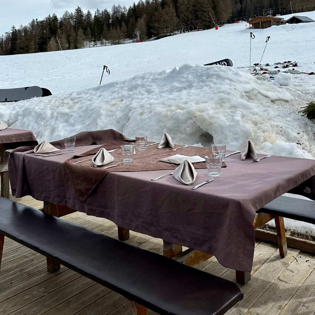 An outdoor dining setup at Chalet du Plan Bois with rustic wooden tables draped in brown tablecloths, set for service against a snowy mountain backdrop.