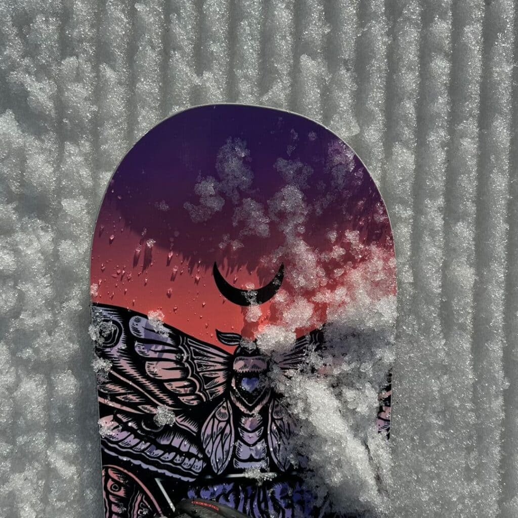 Close-up of a snowboard with a graphic moth and crescent moon design lying on freshly groomed snow, showcasing the creative flair that comes with snowboarding in La Plagne.