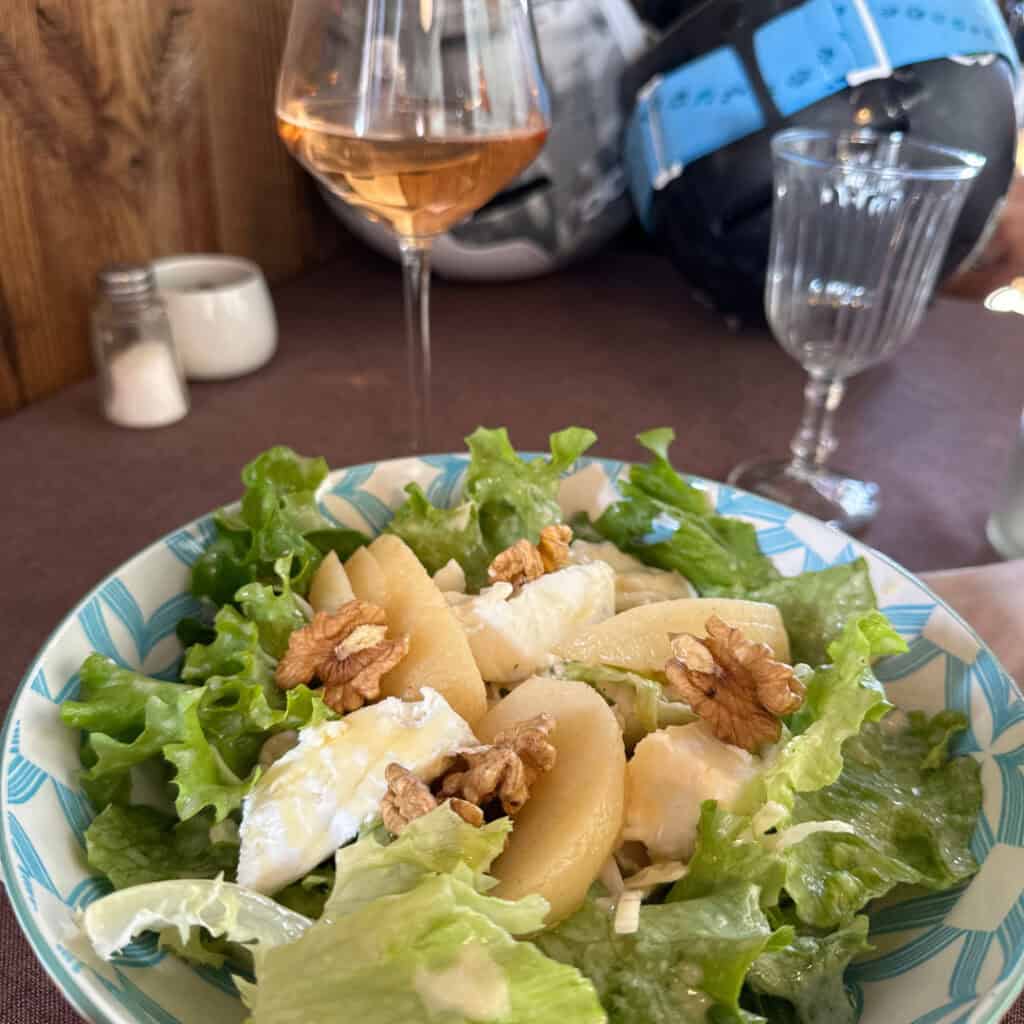 A beautifully plated salad at Chalet du Plan Bois with poached pears, brie cheese, walnuts, and fresh lettuce, served alongside a glass of rosé wine and ski gear in the background.