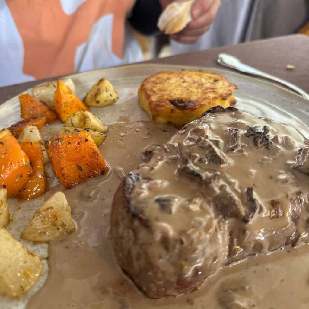 A hearty dish served at Chalet du Plan Bois featuring a filet of beef topped with creamy mushroom sauce, roasted root vegetables, and a golden potato cake.