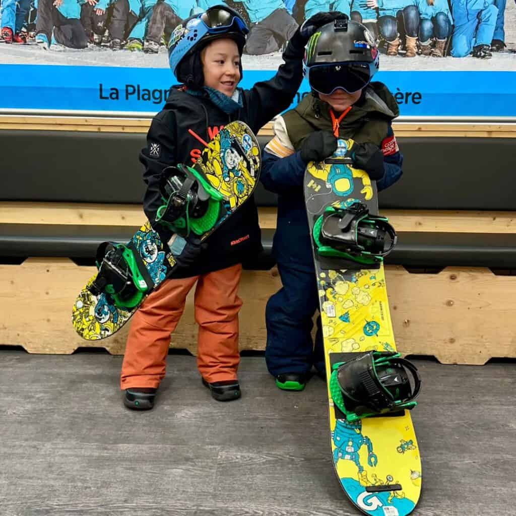Two kids hold brightly colored snowboards with cartoon graphics at an indoor lodge in La Plagne, showing how fun and accessible snowboarding is for young riders at a resort known for skiing too.
