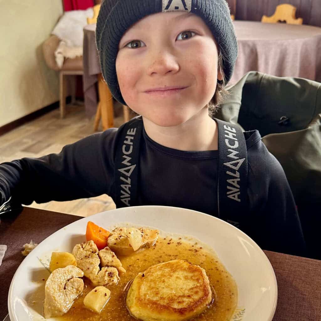A smiling child in ski gear enjoys a warm meal at Chalet du Plan Bois, with tender chicken, root vegetables, and a crispy potato cake in mustard sauce on the plate.