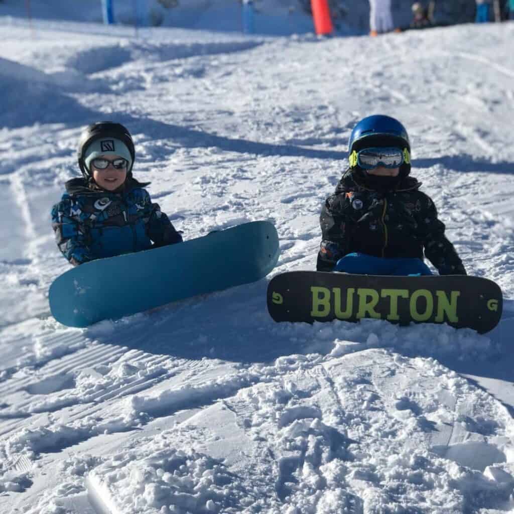 Kids sit with their snowboards on the snow at La Plagne, one with a bold "BURTON" logo, enjoying the slopes—proof that both skiing or snowboarding in La Plagne is a hit with kids.