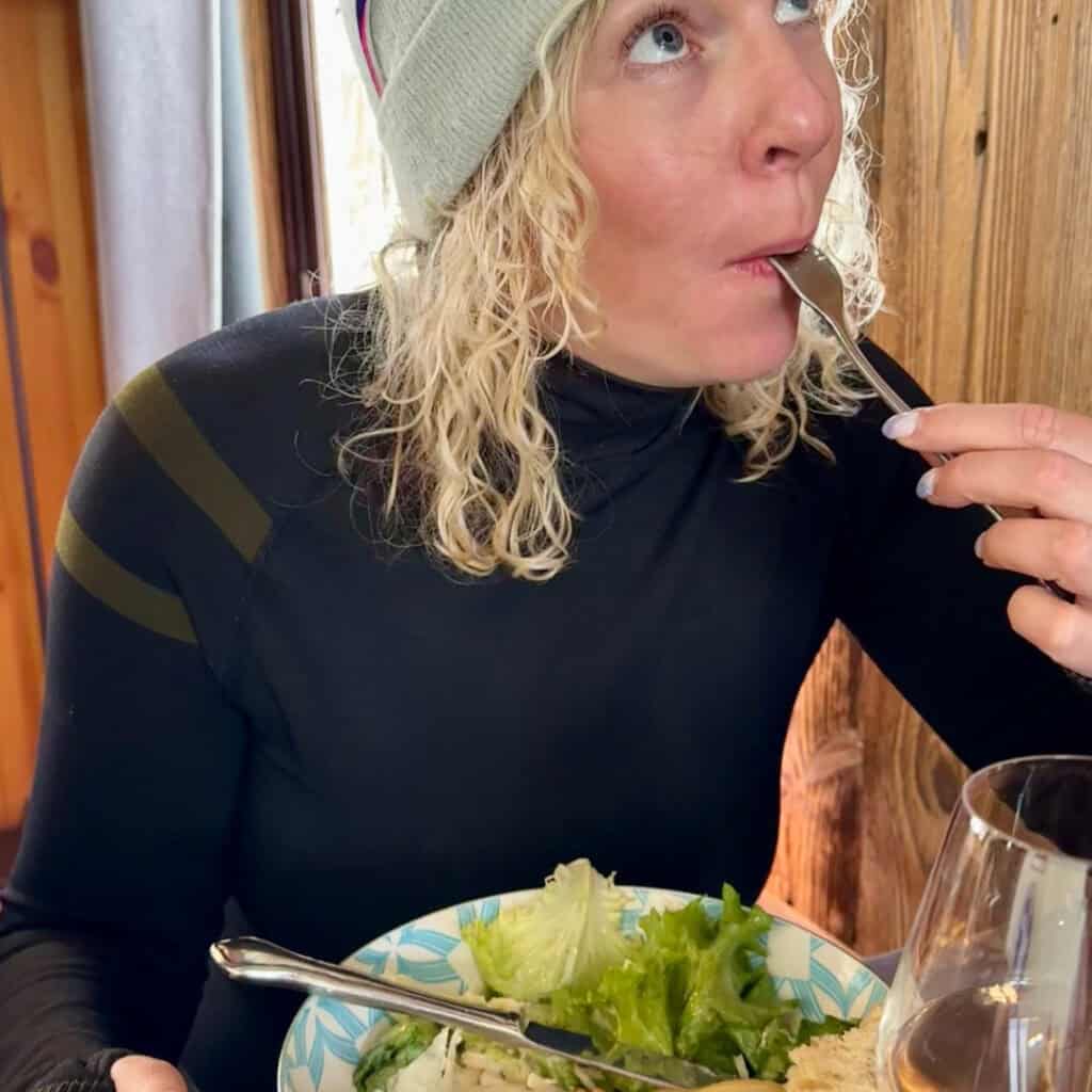 A woman in ski attire takes a bite of her salad at Chalet du Plan Bois, with bright green lettuce and rosé wine visible in the foreground, highlighting the cozy après-ski dining vibe.