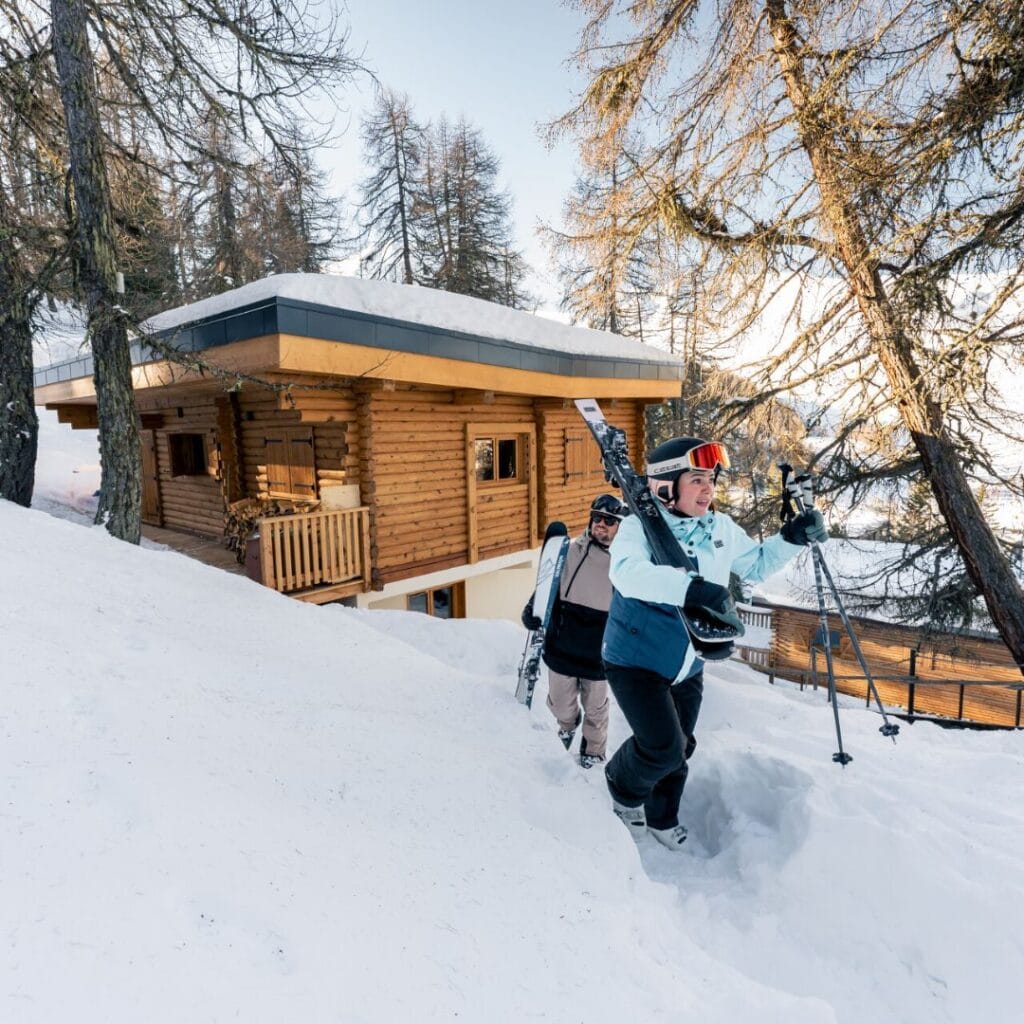 Two skiers walk through deep snow toward a cozy wooden chalet tucked in the trees—an inviting example of the best accommodation Plagne Centre for nature-lovers and ski-in convenience.