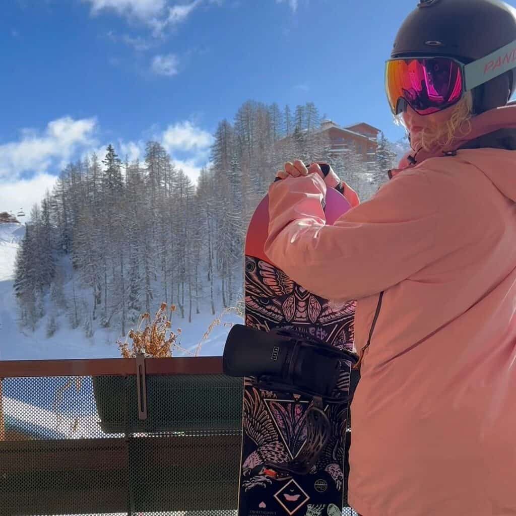 A snowboarder in a pink jacket stands on a snowy balcony overlooking La Plagne's forested slopes, capturing the peaceful side of both skiing or snowboarding in this alpine resort.