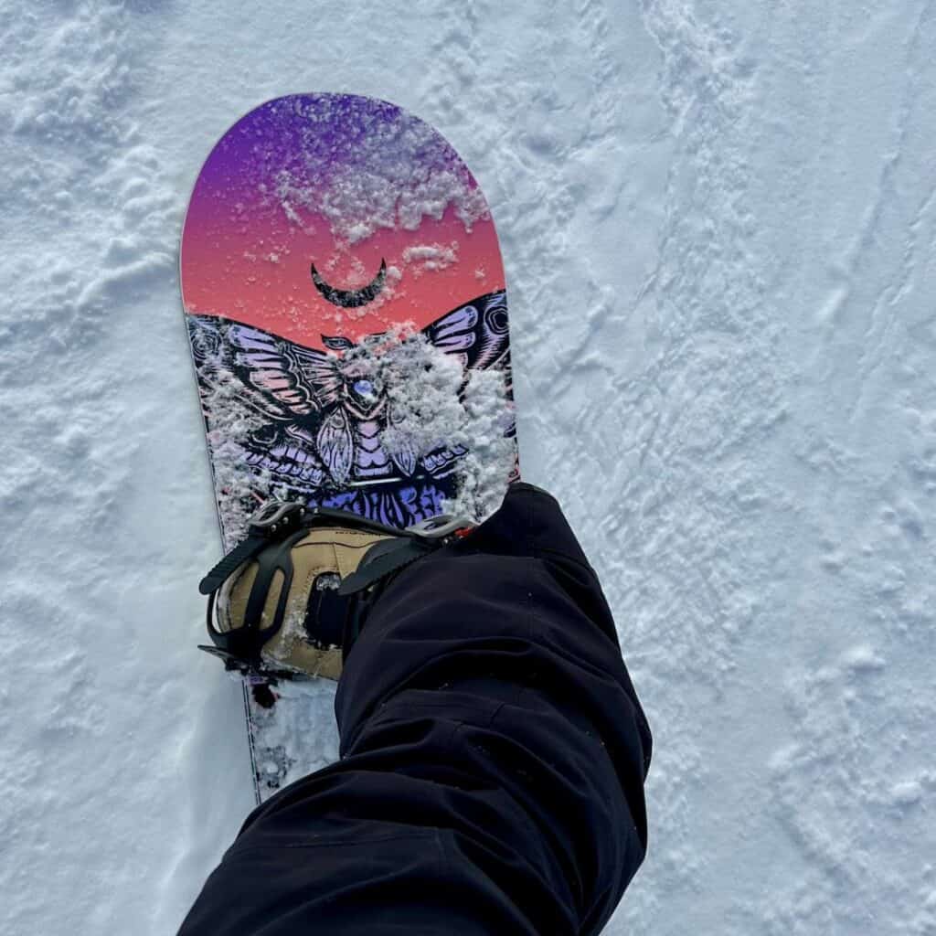 View of a snowboarder’s foot strapped into a crescent moon-themed board against the snowy ground, offering a personal look into the experience of snowboarding in La Plagne.