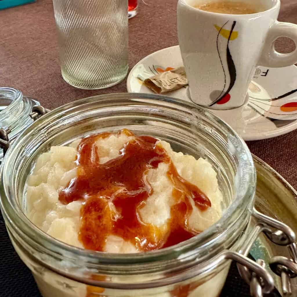A dessert served at Chalet du Plan Bois featuring creamy rice pudding drizzled with caramel sauce in a glass jar, paired with a shot of espresso on a rustic table.