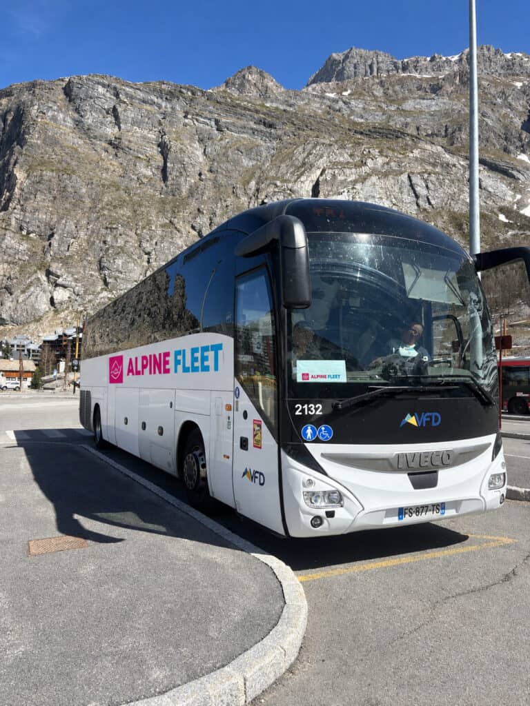White Alpine Fleet branded airport transfer coach parked at a mountain bus stop in the French Alps