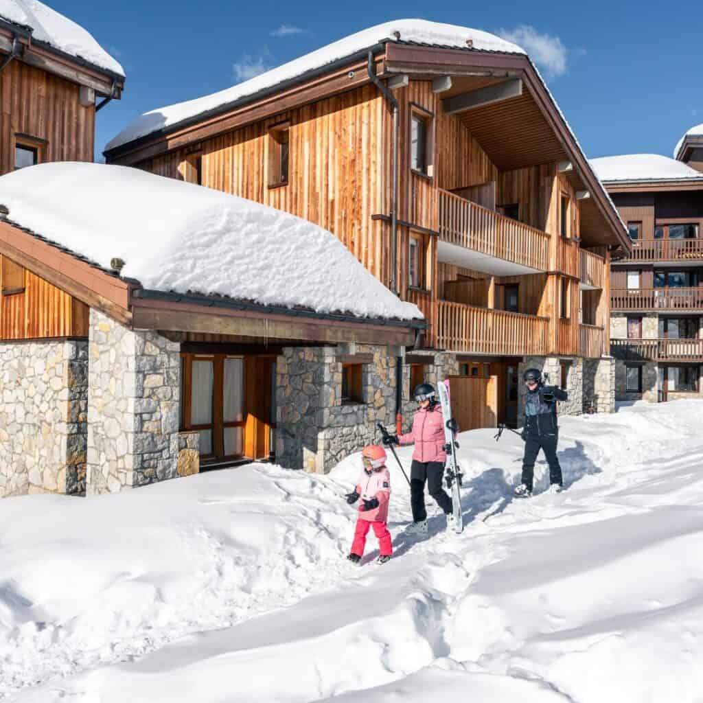 A family dressed in ski gear walks through fresh snow outside a cosy wooden chalet with stone accents, under bright blue skies in Belle Plagne. This inviting alpine lodge, surrounded by snow-covered paths, highlights the best accommodation Belle Plagne has to offer for a winter getaway.