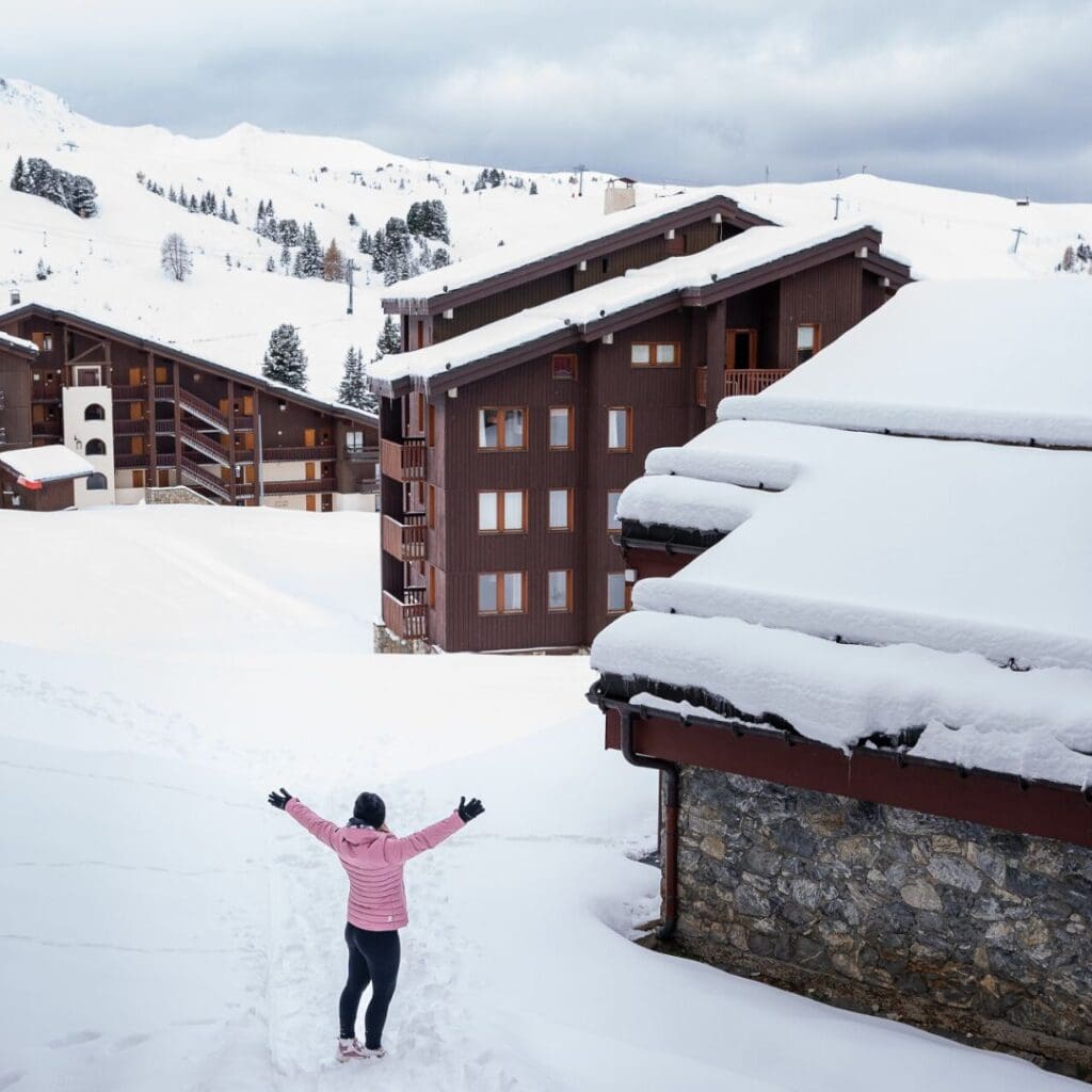 A woman raises her arms in joy as she walks through deep snow in a Belle Plagne village, surrounded by ski-in/ski-out lodges considered among the best accommodation Belle Plagne options.