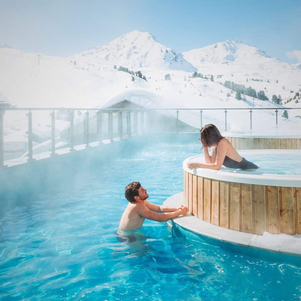 A couple relaxes in a steaming outdoor pool at the Eeep Nature Spa in Belle Plagne, surrounded by snow and mountain views.