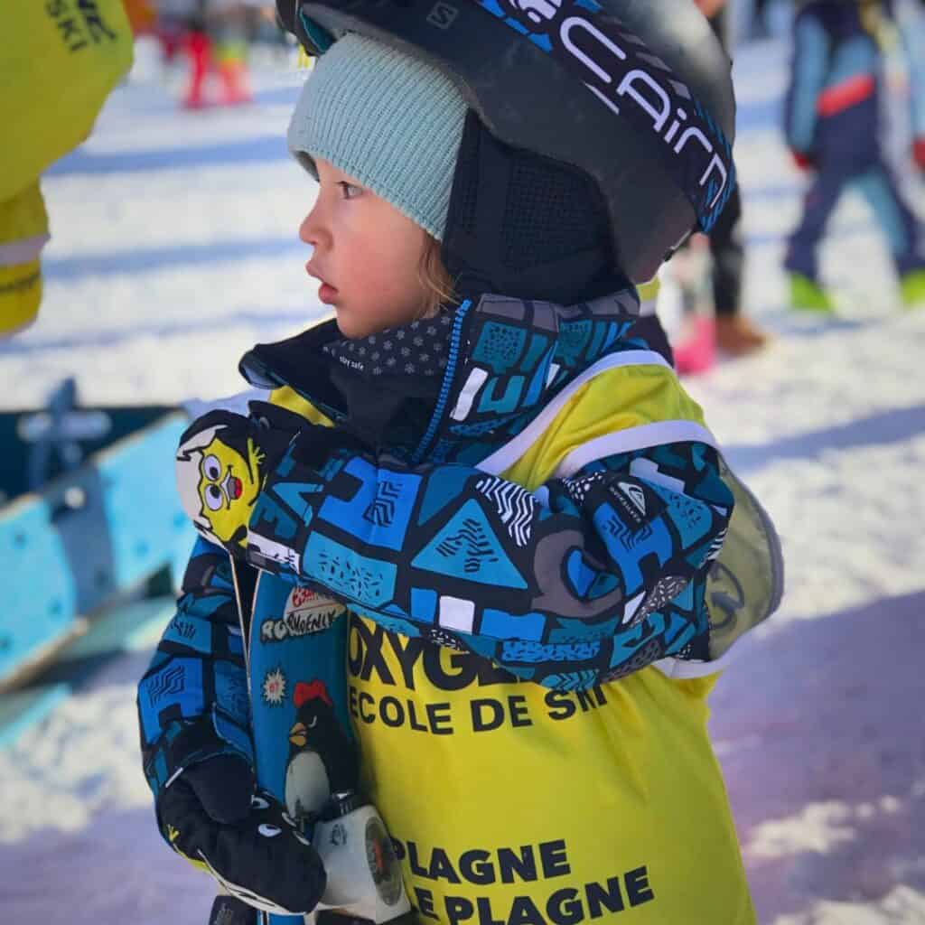 A young child in colorful ski gear and a yellow Oxygène ski school vest prepares for a lesson, highlighting family-friendly services near the best accommodation Belle Plagne.