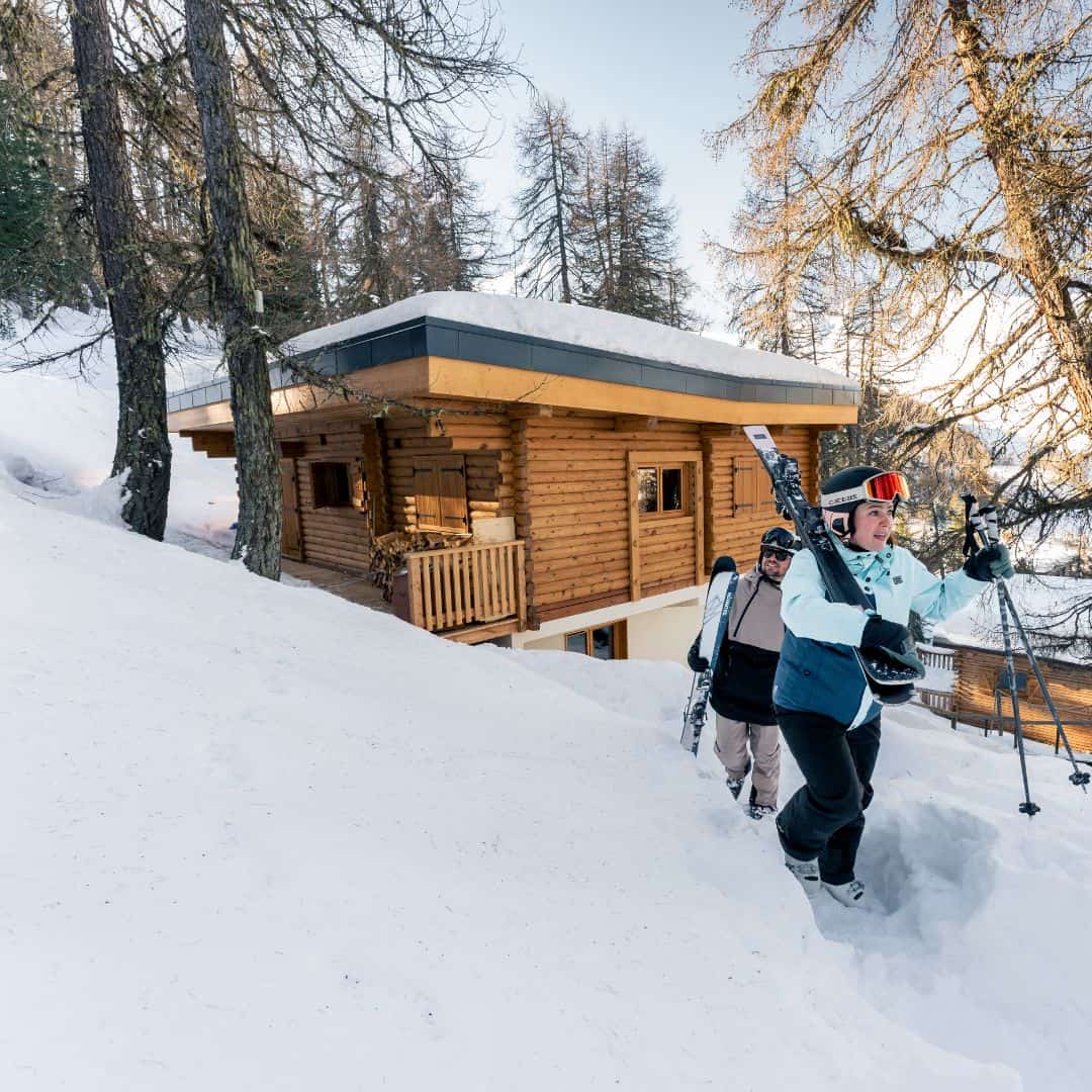 Two skiers in full gear walk uphill through snow toward a cozy wooden chalet surrounded by pine trees in La Plagne Centre. The cabin's modern-rustic design and prime slope-side location highlight it as an ideal ski-in, ski-out accommodation.