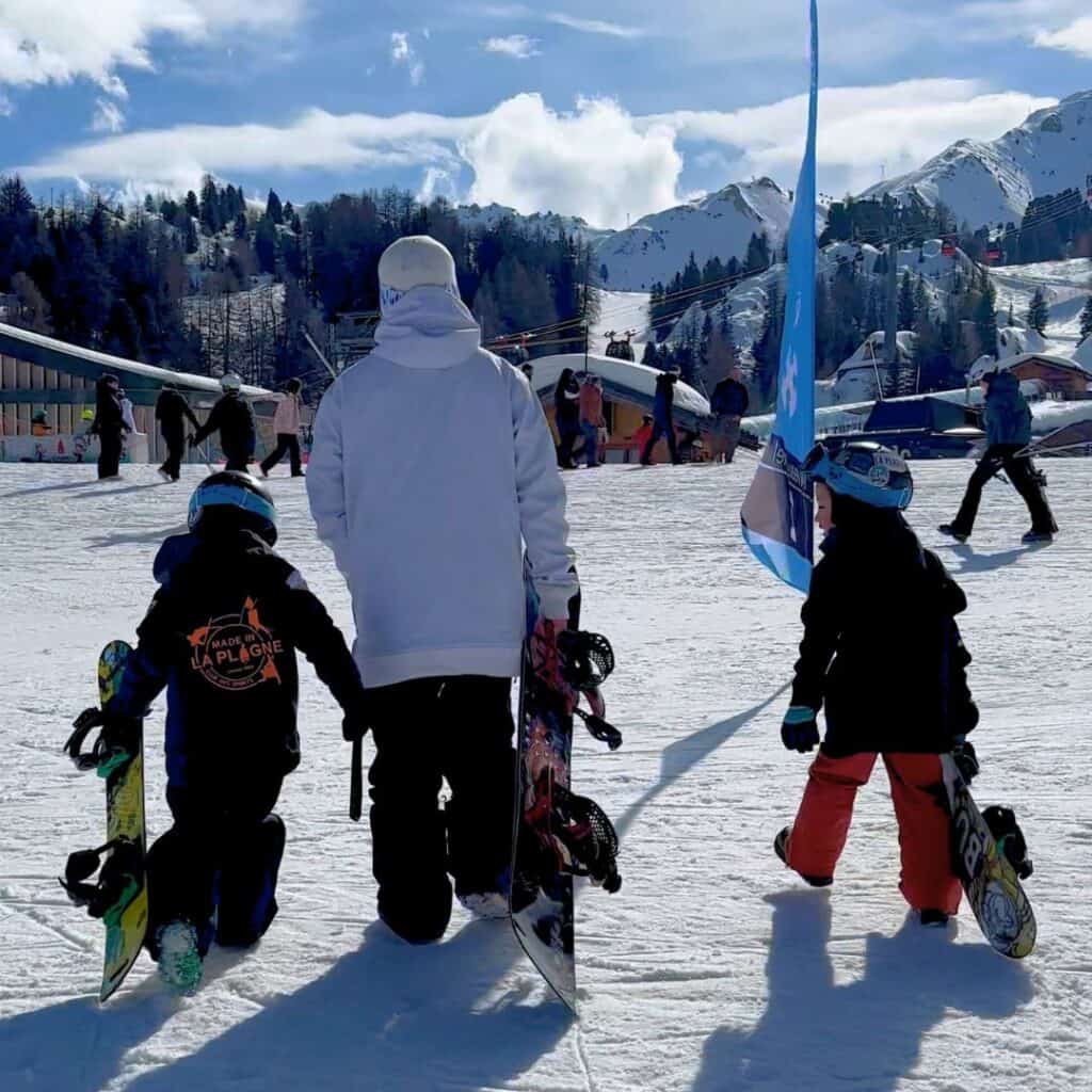 A parent and two kids walk on a snowy slope in La Plagne, each carrying a snowboard. The kids are dressed in full kids snowboarding gear, including helmets, snow pants, gloves, and jackets, with one jacket reading “Made in La Plagne.” Snow-covered mountains and ski lifts are visible in the background.