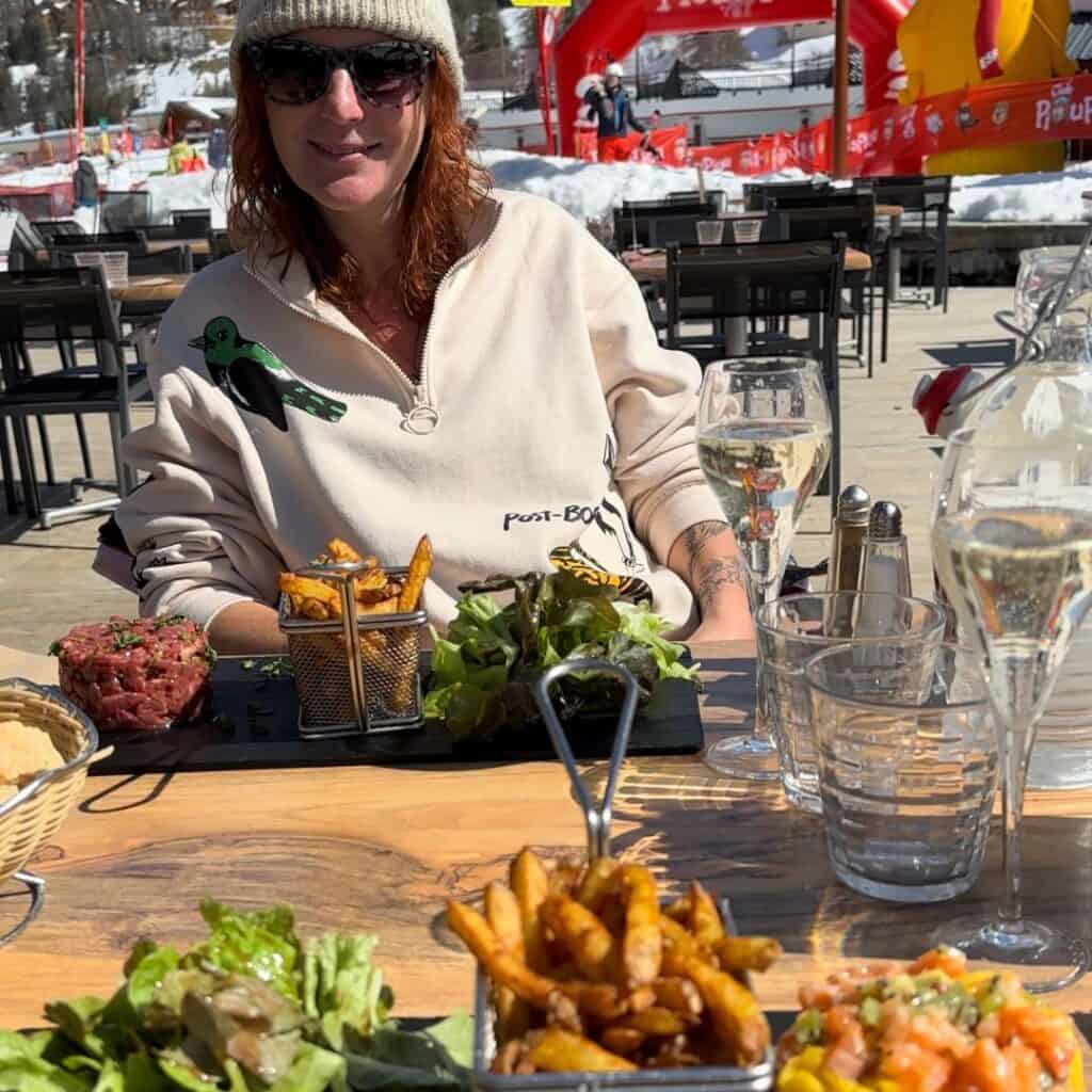A woman enjoys steak tartare, fries, and champagne on a sunny terrace in Plagne Centre—just steps away from the best accommodation Plagne Centre for food lovers.