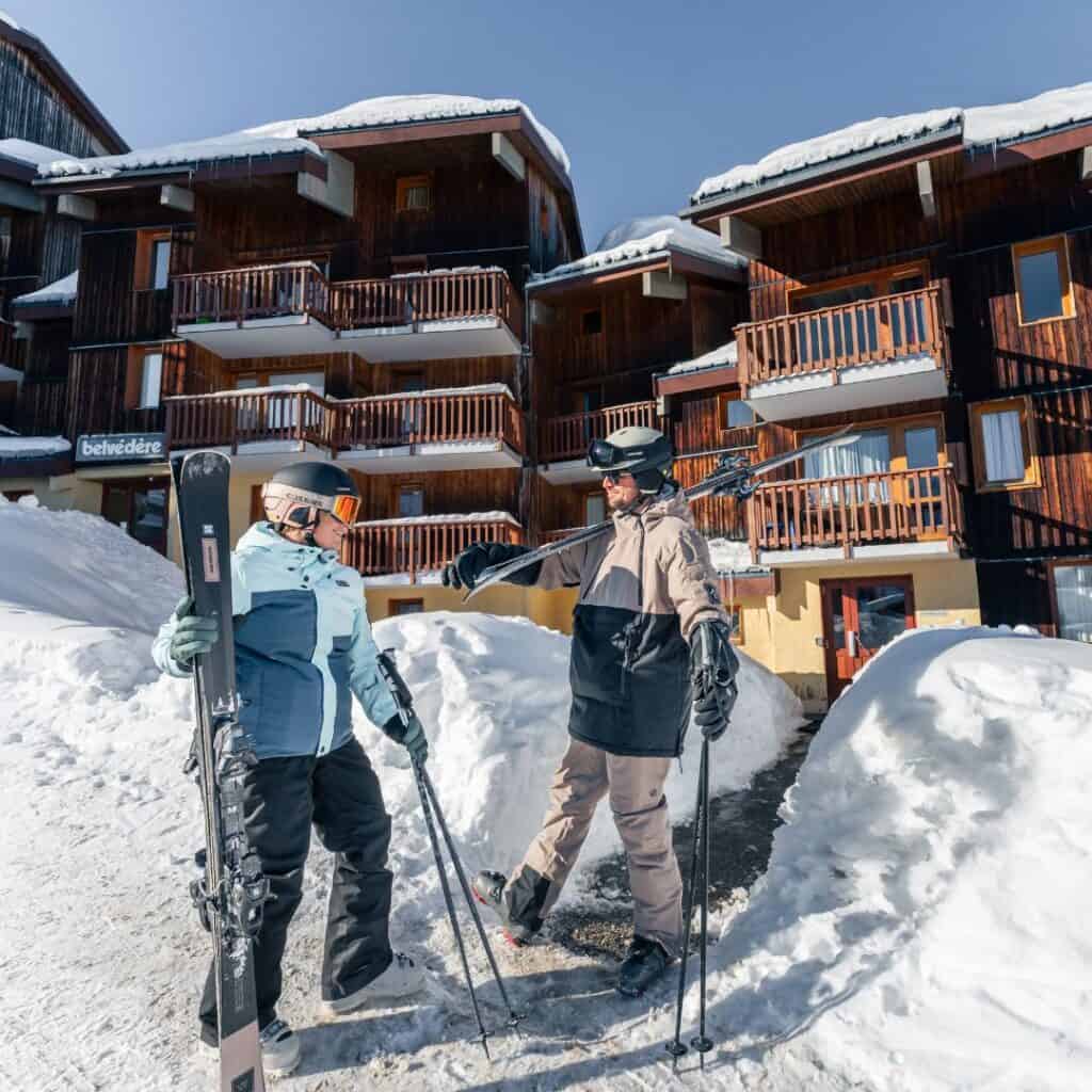 Two skiers stand outside a cozy chalet in La Plagne, chatting and holding their gear in front of the Belvédère building, a popular option for self catering La Plagne holidays, surrounded by snow and clear blue skies.