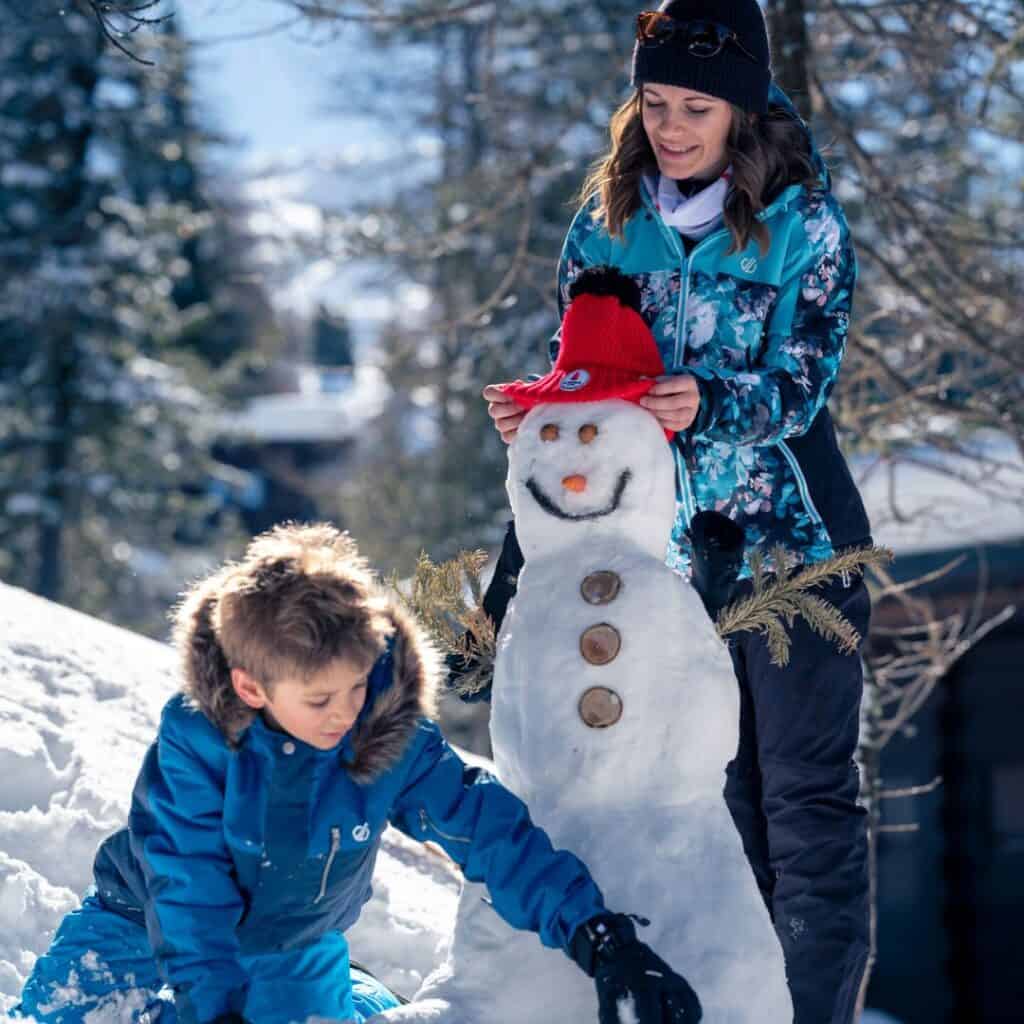 A mother and son build a snowman together outside a self catering La Plagne apartment, adding a red hat and twig arms in a fun family moment.