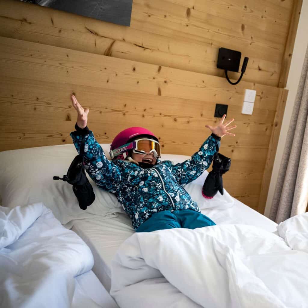 A young child in colorful ski gear and helmet throws their arms up with excitement while lying on a bed inside a cozy self catering La Plagne apartment with wooden walls and warm lighting.