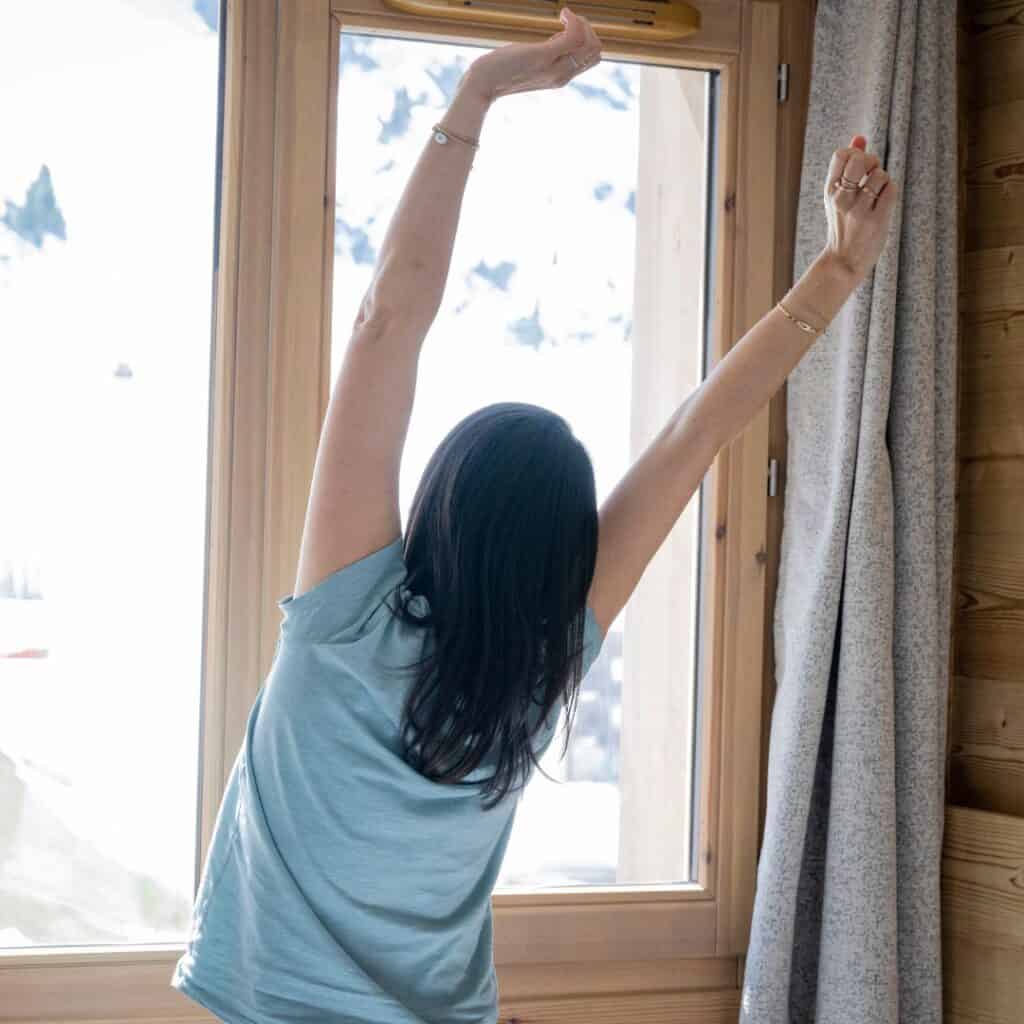 A woman stretches in front of a sunlit window overlooking snowy mountains, enjoying a peaceful morning in a self catering La Plagne chalet.